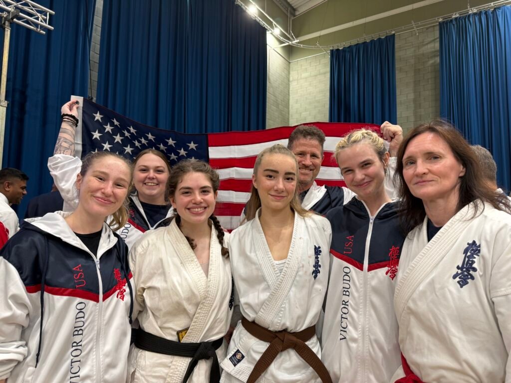 USA-IFK team at the 48th BKK British Open in Crawley, England. Front row, left to right: Senpai Lyric Schenker, Sensei Audrey Bartholomay, Willow Swan, Senpai Caitlyn Anderson, and Elvira Anderson, with Shihan Sean Schenker and other team supporters behind them.